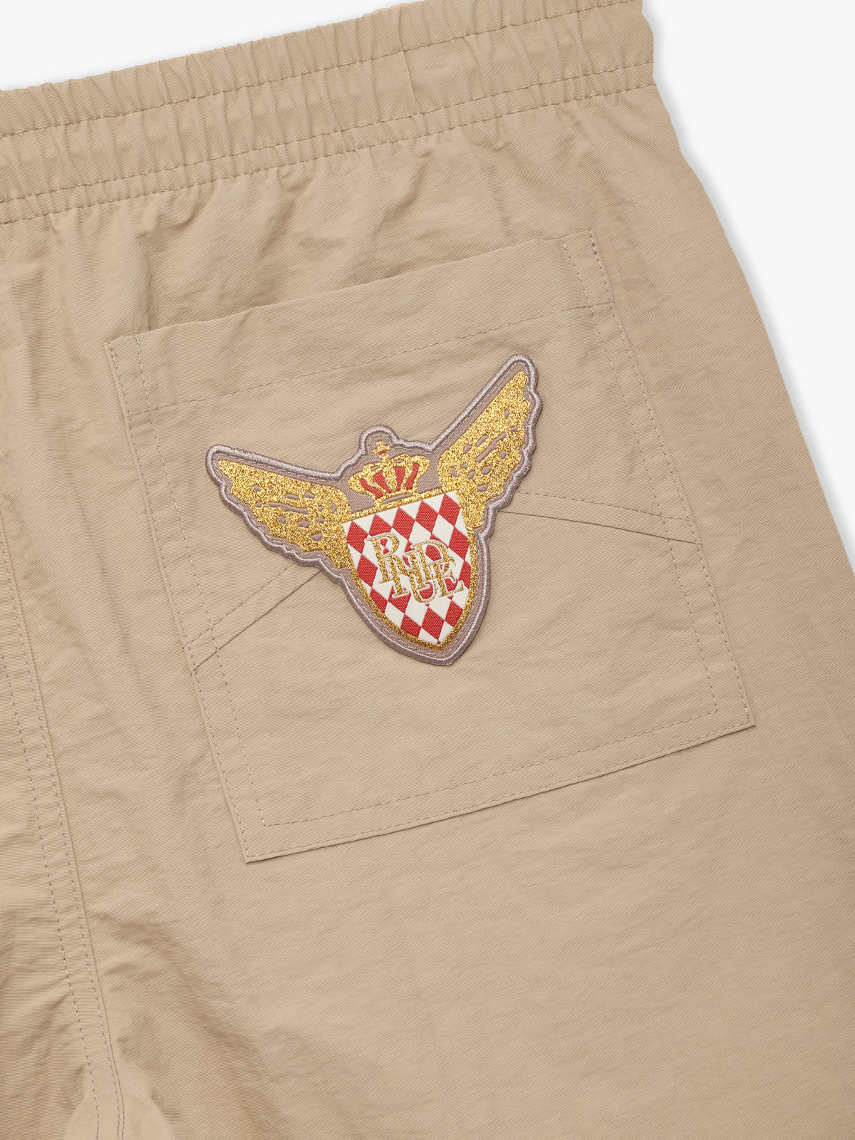 Close-up of the gold winged crest patch with a red and white checkered shield on the rear pocket of the tan shorts