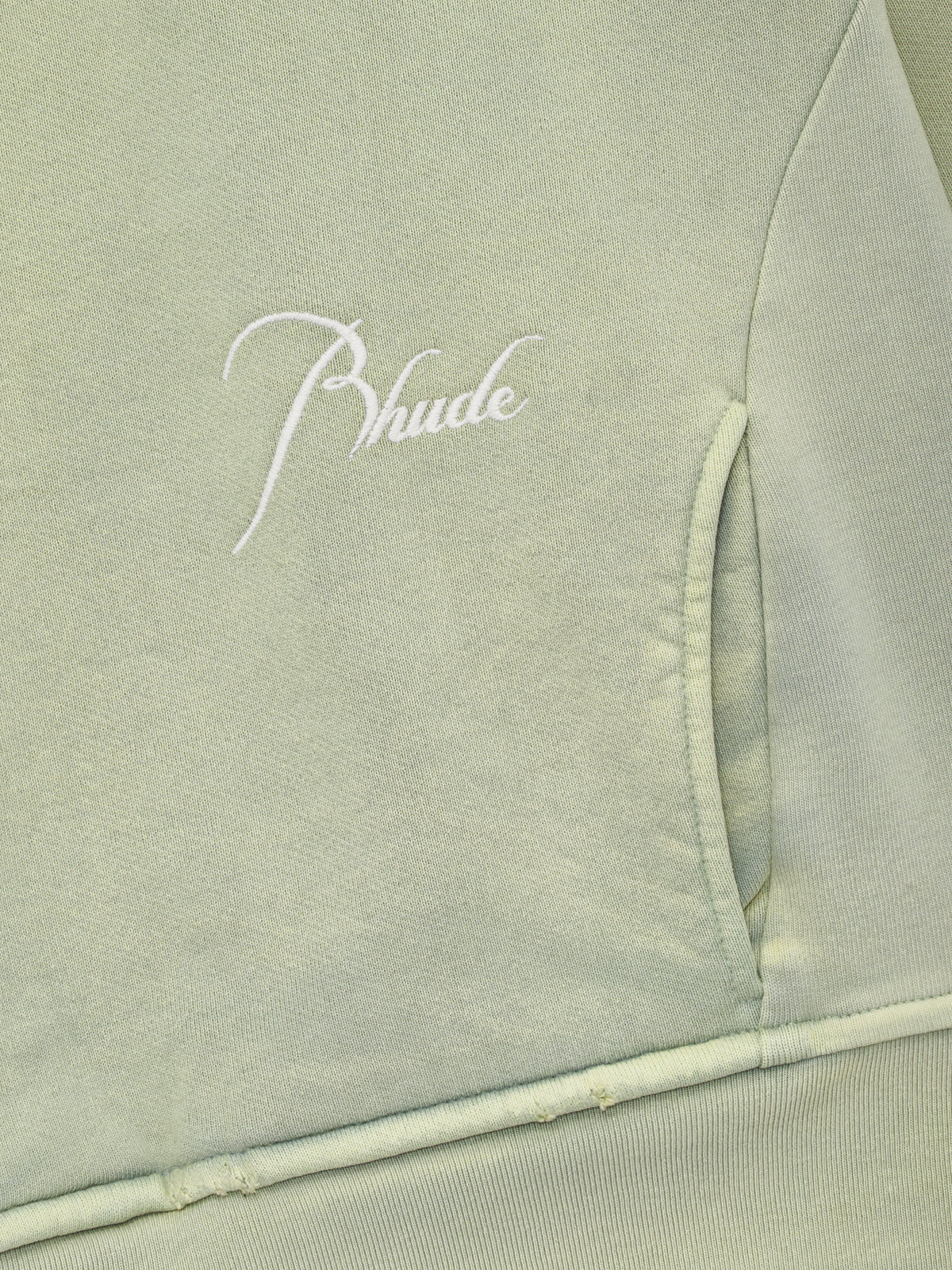 Close-up of the white script Rhude embroidery above the front pocket on the washed seafoam quarter zip