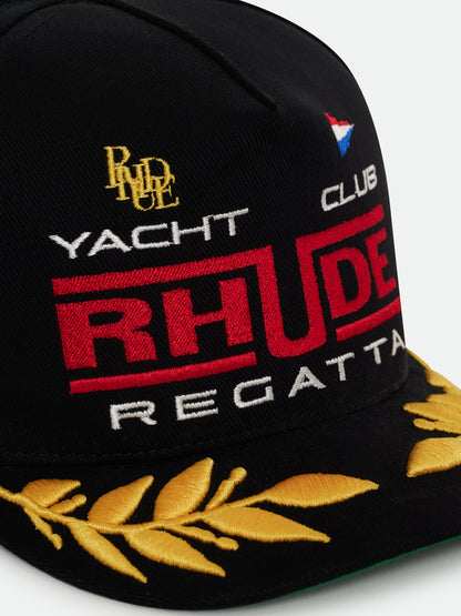 Close-up of the gold RHUDE monogram, pennant flag, red RHUDE “Regatta” lettering and gold laurels on the black cap