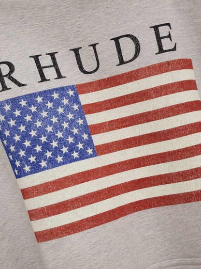 Angled close-up of the cracked US flag print and black RHUDE lettering on the heather grey hoodie
