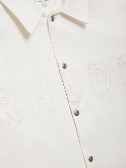 Angled close-up of the raised tonal RHUDE lettering beside the silver snaps on the white coaches jacket