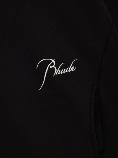 Close-up of the cream script Rhude embroidery beside the curved seam on the vintage black crewneck