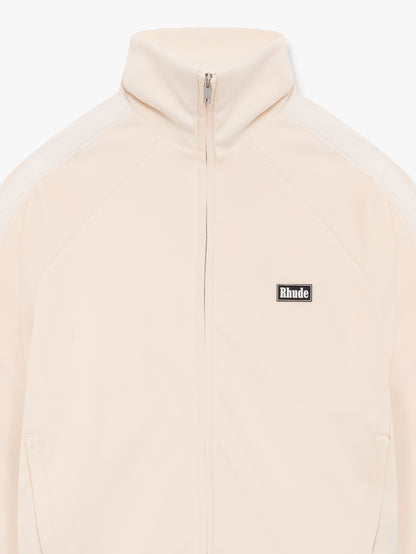 Close-up of the stand collar, zip and black Rhude chest badge on the cream track jacket