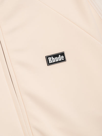Angled close-up of the black and white Rhude badge on the cream track jacket