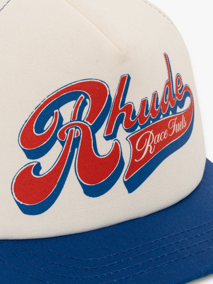 Close-up of the red and blue script Rhude “Race Fuels” print on the cream front panel above the blue brim