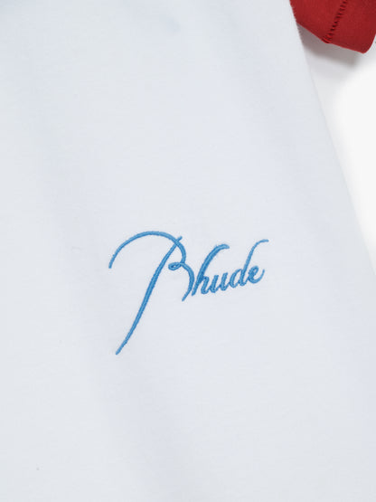 Close-up of the small sky blue script Rhude embroidery near the hem of the white and red raglan tee