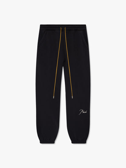 Black cuffed sweatpants with long tan drawcords and cream script Rhude embroidery on the lower leg, front view