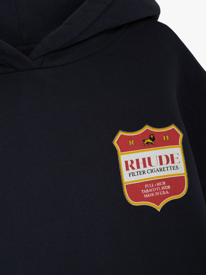Angled close-up of the red and gold RHUDE crest on the chest of the vintage black hoodie