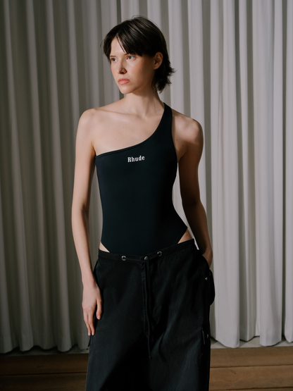Model wearing the washed black parachute pants low on the hips with a black one-shoulder Rhude bodysuit, three-quarter view