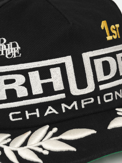 Close-up of the raised white RHUDE “Champions” lettering, gold “1ST” and laurel embroidery on the black cap