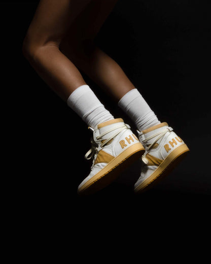 Model wearing the white and mustard high-top sneakers with white socks, both feet in the air against a black background